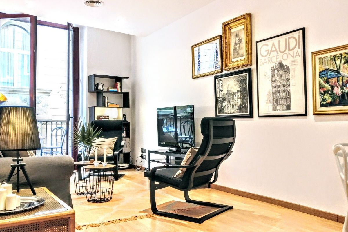 this common area boasts comfortable sofa, two relaxing armchairs, designer tripod lamps and a convenient bookcase with books, for any relaxing moment at this flat for rent in Barcelona central location furnished apartment 